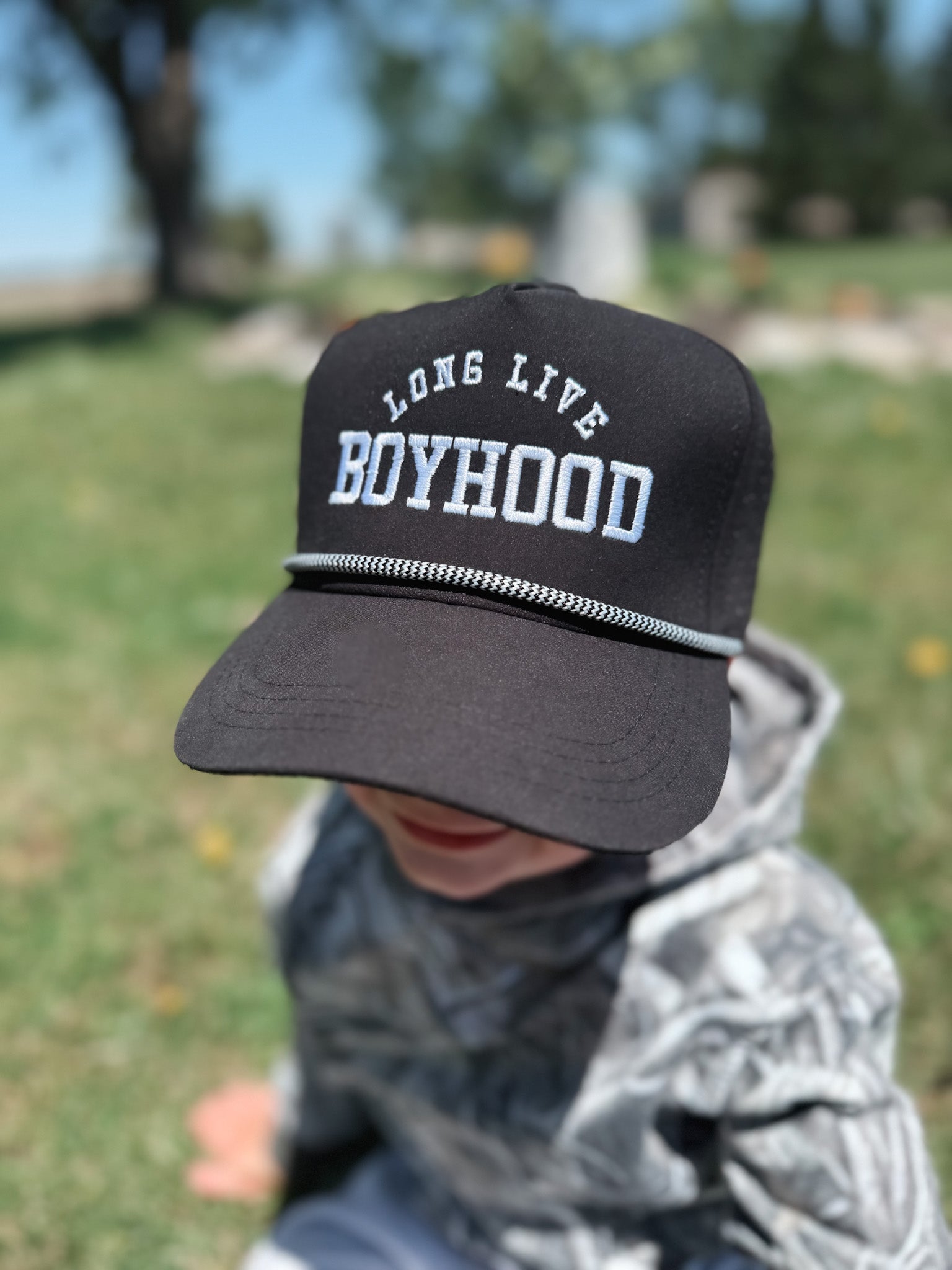 Child wearing a black cap with 'Long Live Boyhood' text in a park setting