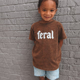 Child wearing a brown t-shirt with 'feral' printed on it against a gray brick wall.
