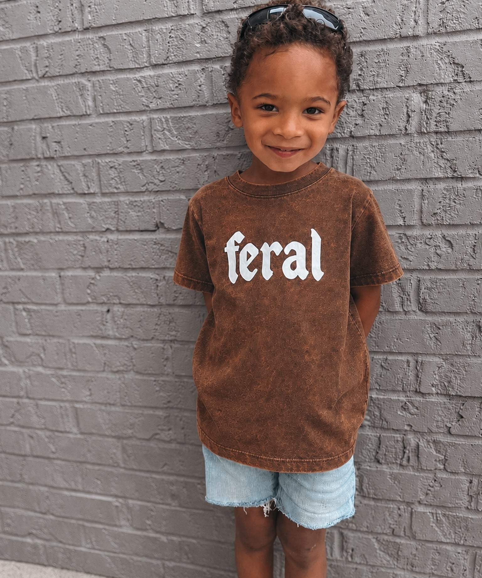 Child wearing a brown t-shirt with 'feral' printed on it against a gray brick wall.