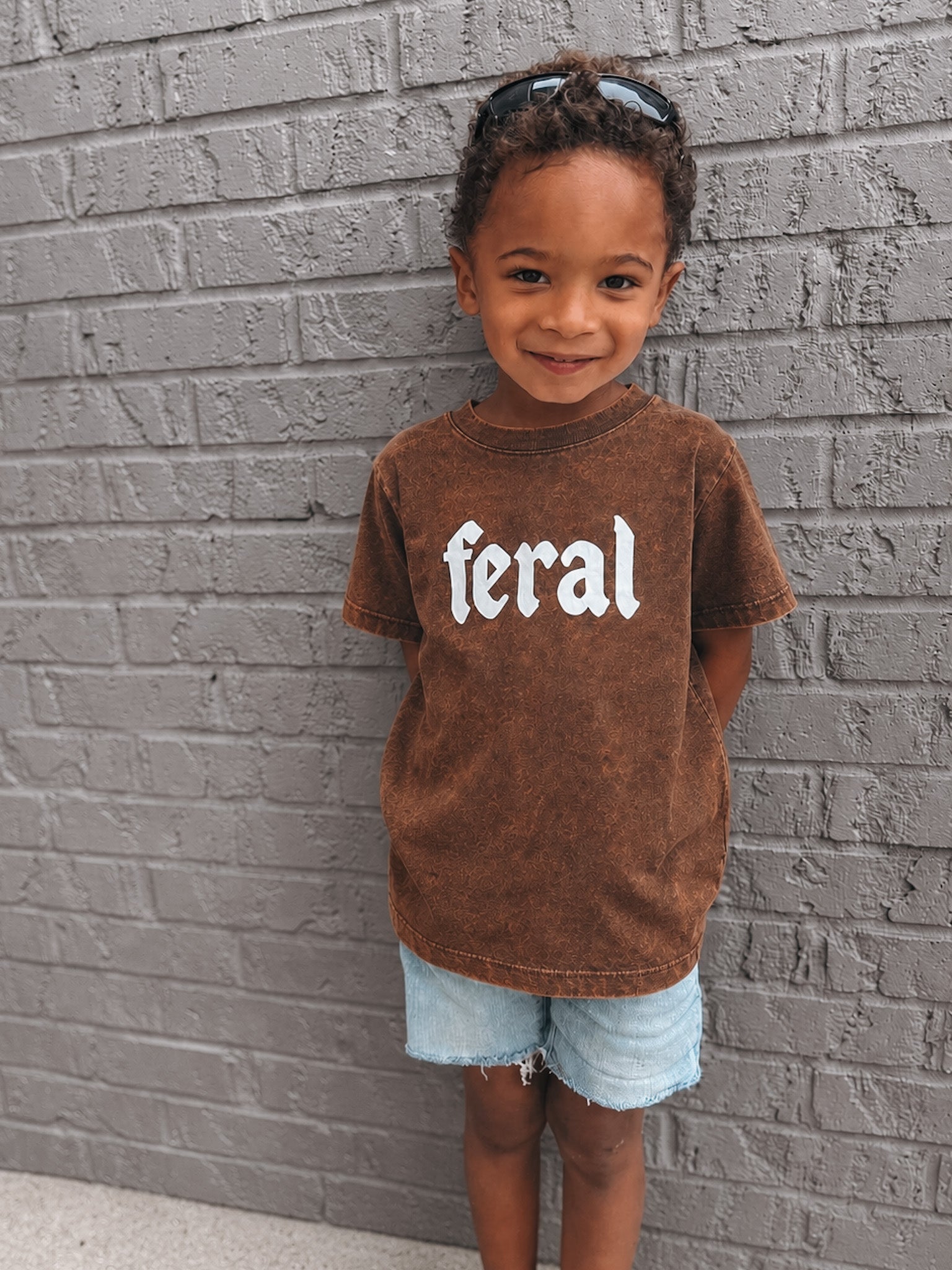 Child wearing a brown t-shirt with 'feral' printed on it against a gray brick wall.