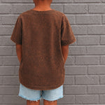 Child wearing a brown t-shirt and denim shorts standing against a gray brick wall.