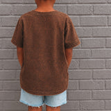 Child wearing a brown t-shirt and denim shorts standing against a gray brick wall.