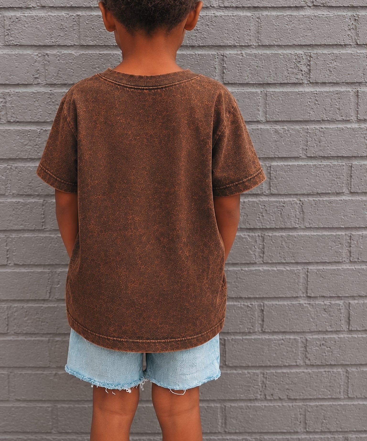 Child wearing a brown t-shirt and denim shorts standing against a gray brick wall.