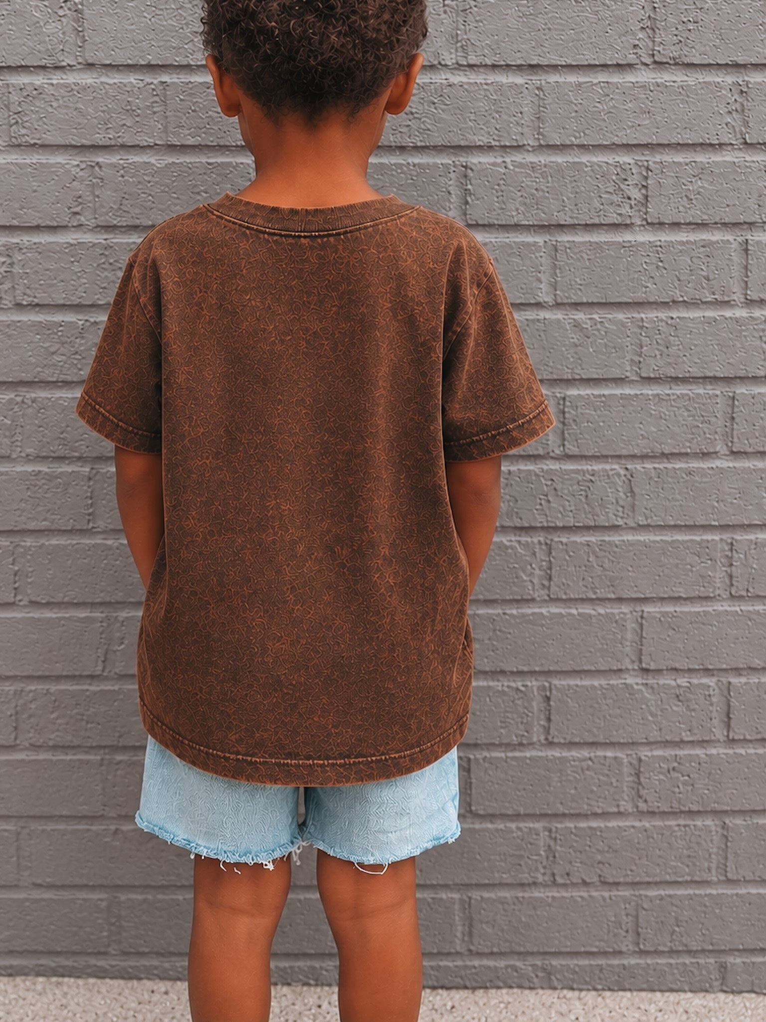 Child wearing a brown t-shirt and denim shorts standing against a gray brick wall.