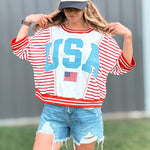 Person wearing a red, white, and blue 'USA' sweatshirt with denim shorts and a cap.