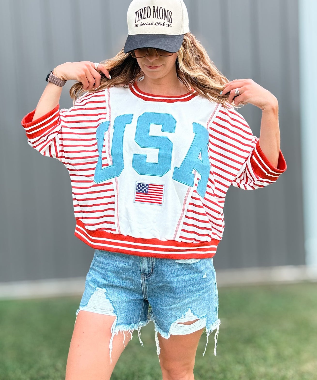 Person wearing a red, white, and blue 'USA' sweatshirt with denim shorts and a cap.