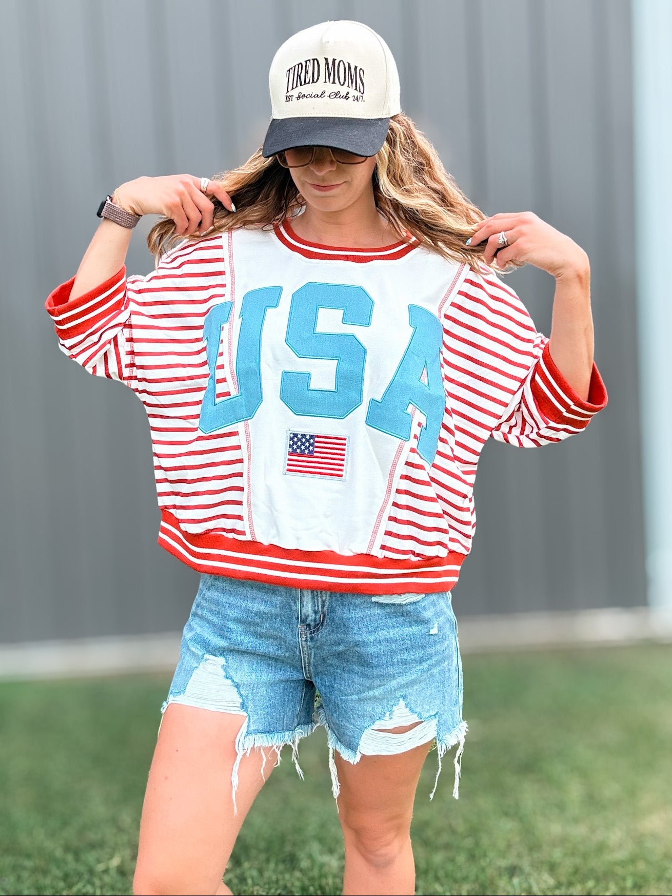 Person wearing a red, white, and blue 'USA' sweatshirt with denim shorts and a cap.