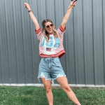 Woman in a red, white, and blue shirt with a graphic design, denim shorts, and white sandals, standing outdoors with arms raised.