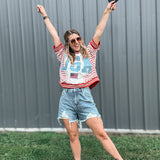 Woman in a red, white, and blue shirt with a graphic design, denim shorts, and white sandals, standing outdoors with arms raised.
