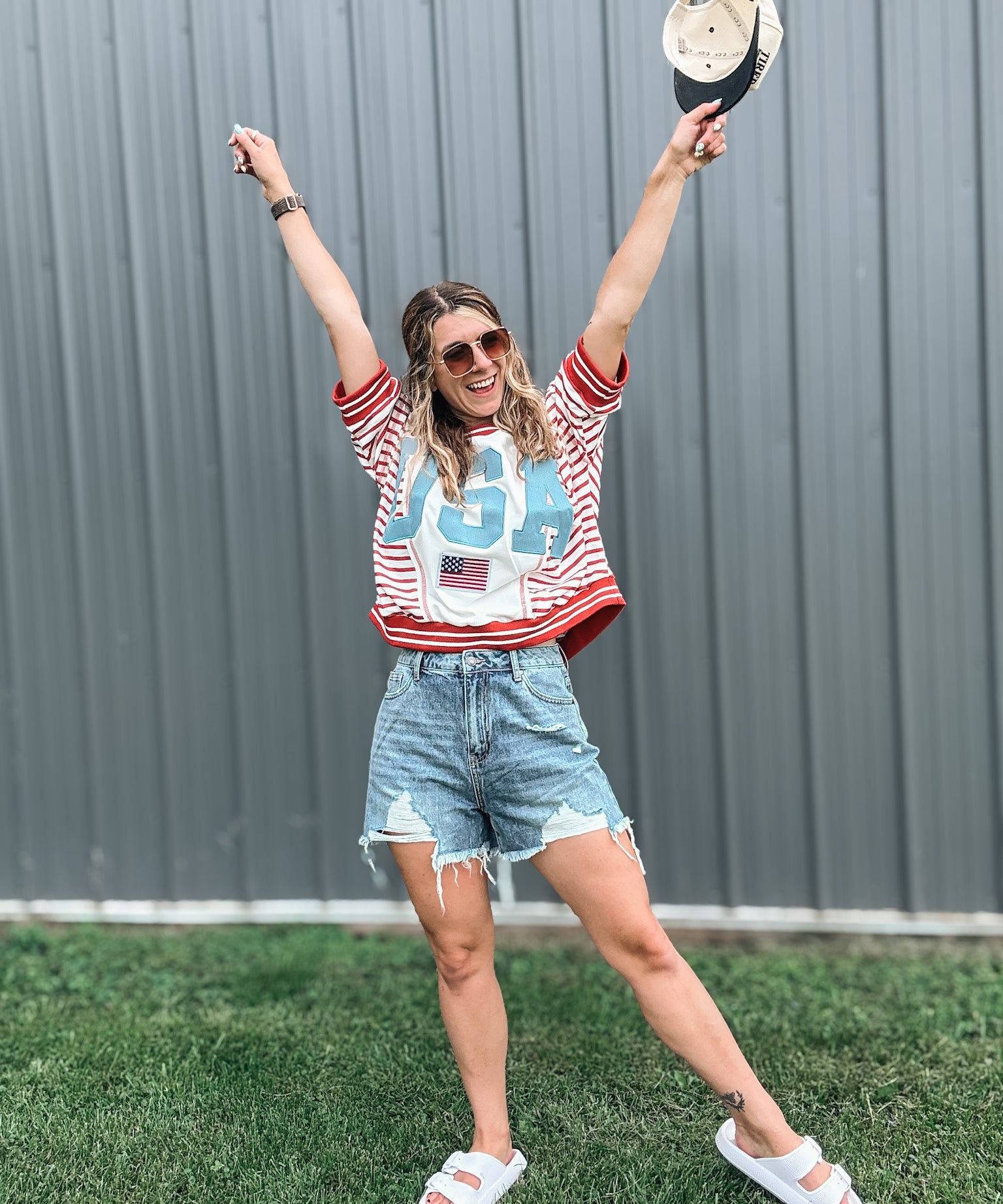 Woman in a red, white, and blue shirt with a graphic design, denim shorts, and white sandals, standing outdoors with arms raised.