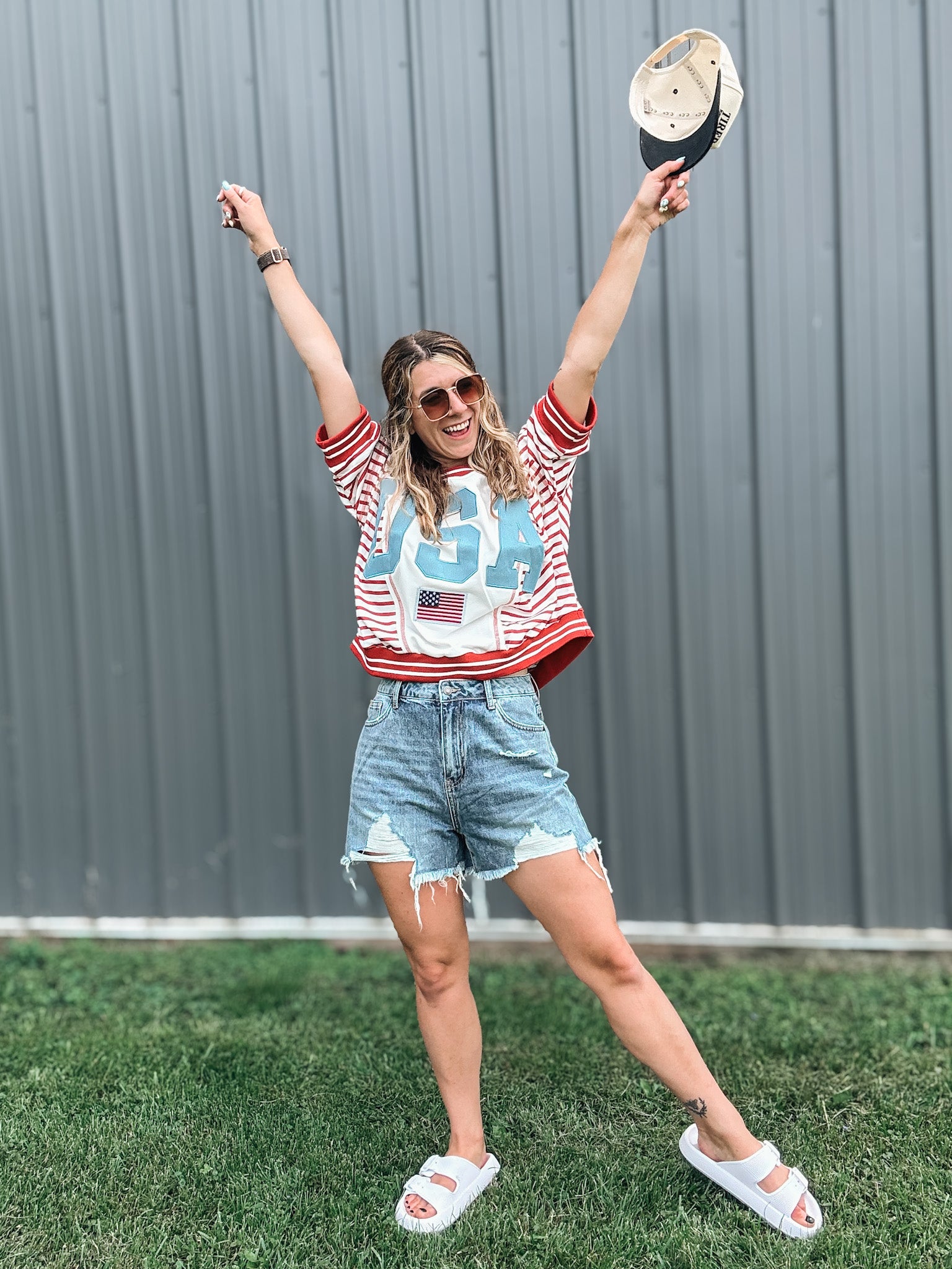 Woman in a red, white, and blue shirt with a graphic design, denim shorts, and white sandals, standing outdoors with arms raised.