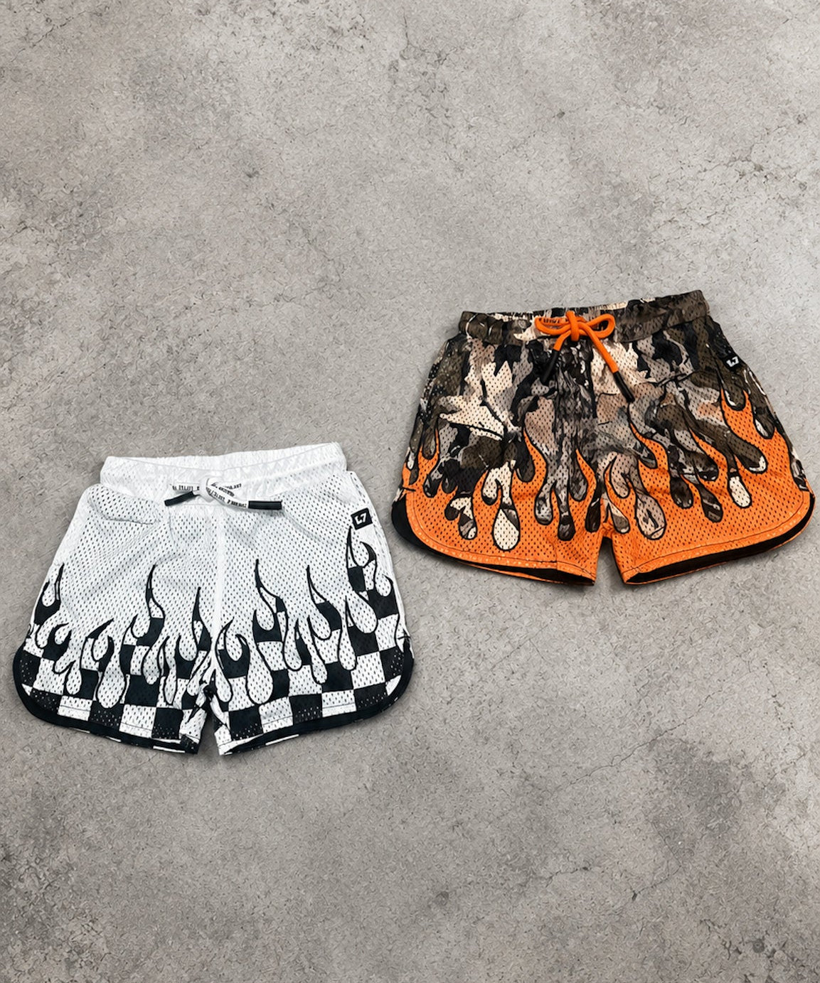 Two pairs of shorts, one white with black flame design and the other orange with black pattern, on a gray concrete surface.