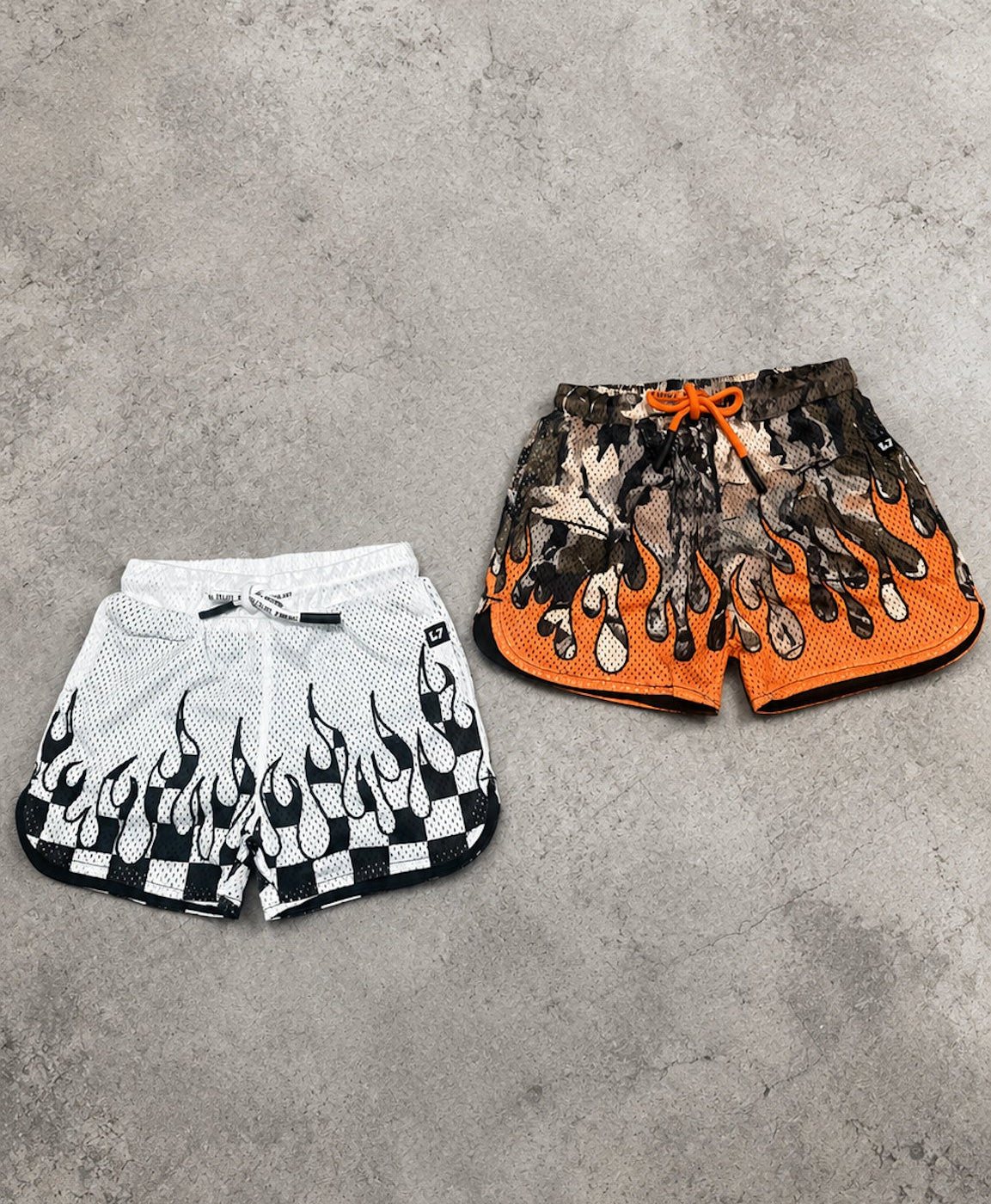 Two pairs of shorts, one white with black flame design and the other orange with black pattern, on a gray concrete surface.
