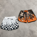 Two pairs of shorts, one white with black flame design and the other orange with black pattern, on a gray concrete surface.