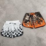 Two pairs of shorts, one white with black flame design and the other orange with black pattern, on a gray concrete surface.