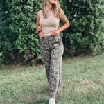Woman in camouflage pants and a beige top standing in a grassy area with greenery.