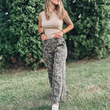 Woman in camouflage pants and a beige top standing in a grassy area with greenery.
