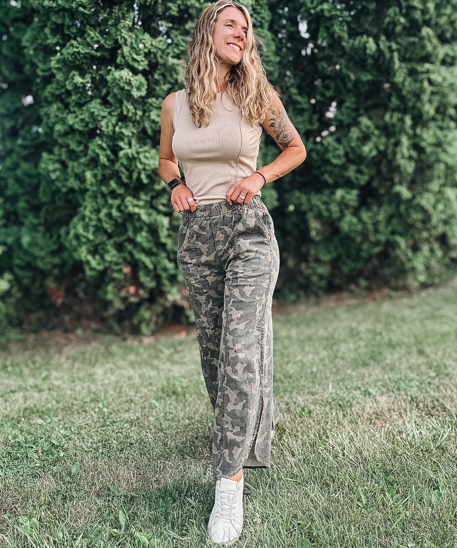 Woman in camouflage pants and a beige top standing in a grassy area with greenery.