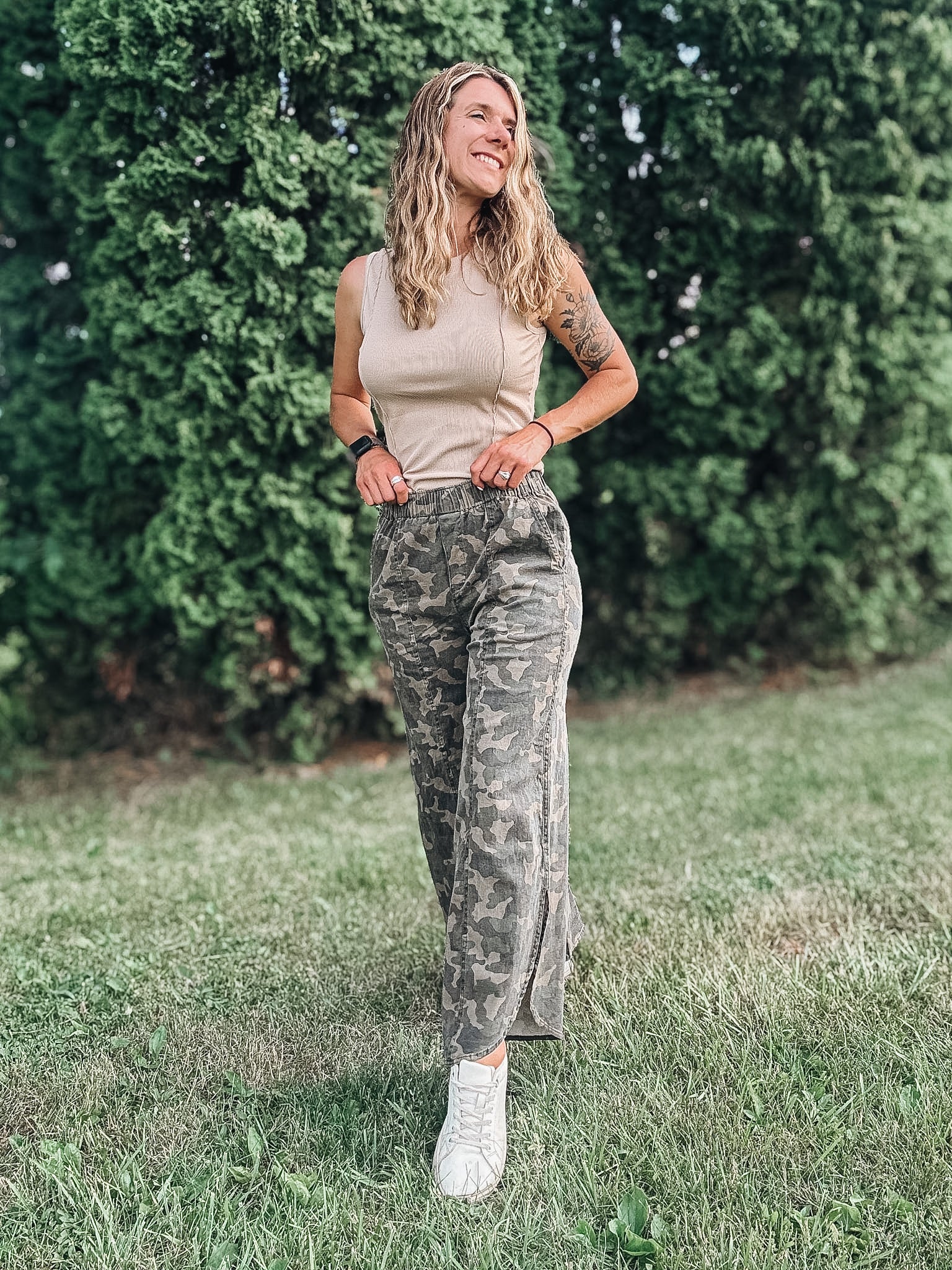 Woman in camouflage pants and a beige top standing in a grassy area with greenery.