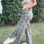 Person wearing camouflage pants and a beige top standing outdoors with greenery in the background
