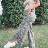 Person wearing camouflage pants and a beige top standing outdoors with greenery in the background