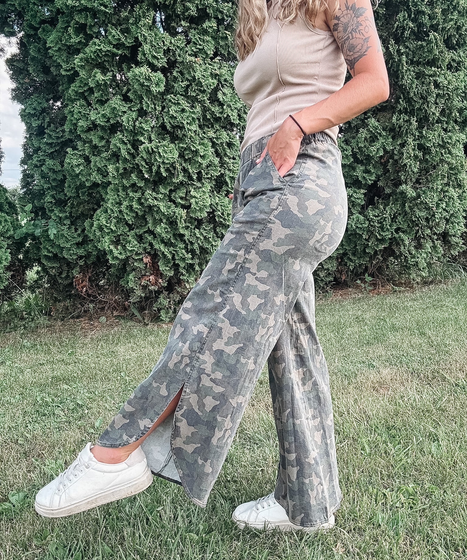 Person wearing camouflage pants and a beige top standing outdoors with greenery in the background