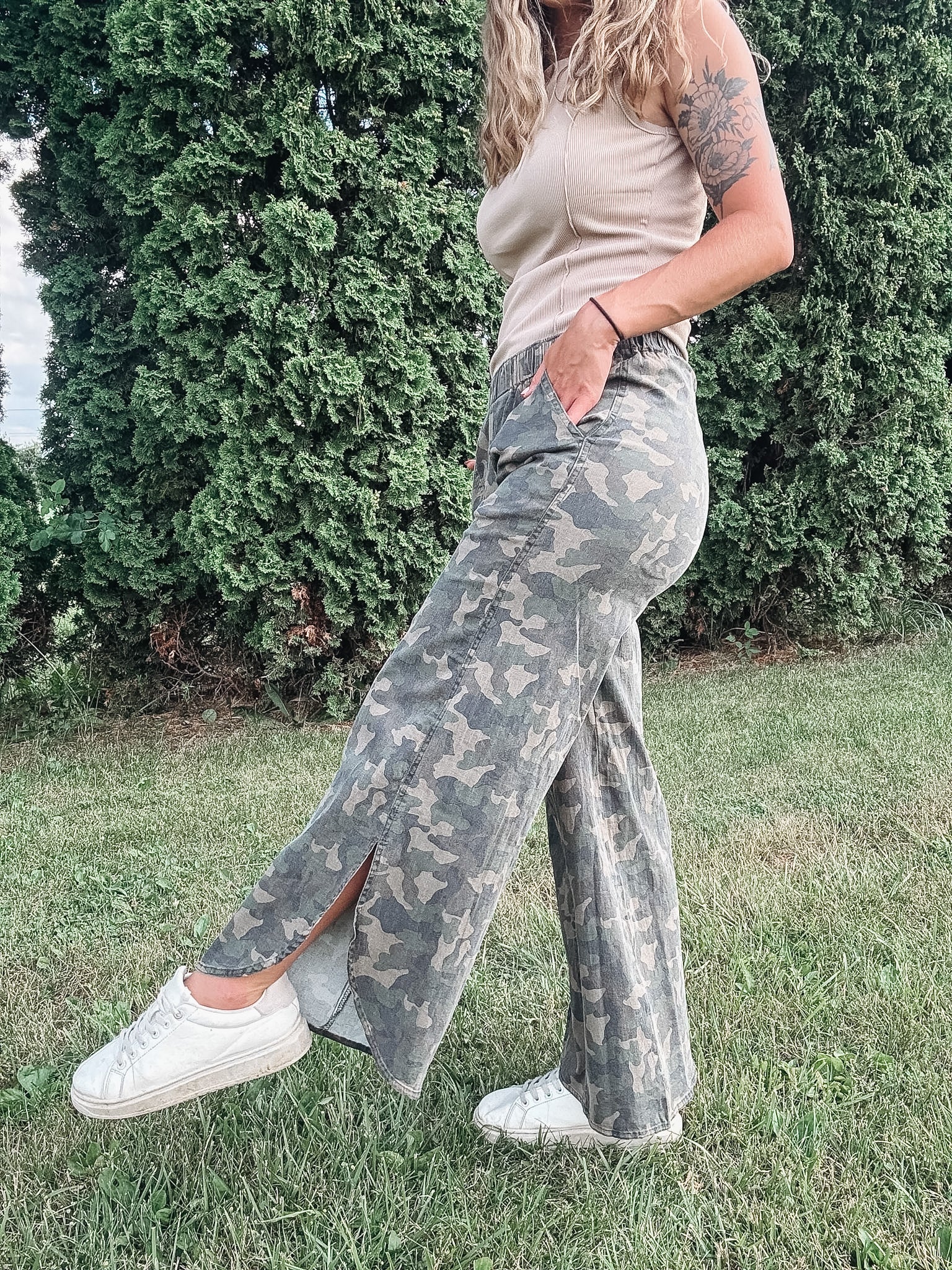 Person wearing camouflage pants and a beige top standing outdoors with greenery in the background