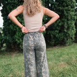 Person wearing camouflage pants and a beige top standing in a grassy area with trees in the background