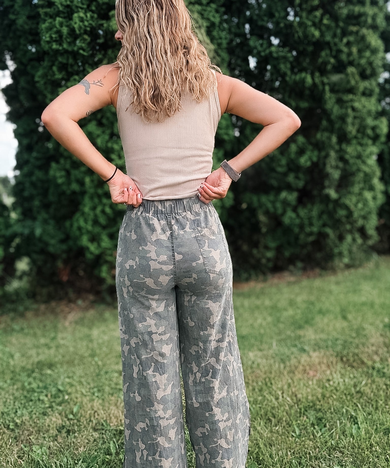 Person wearing camouflage pants and a beige top standing in a grassy area with trees in the background