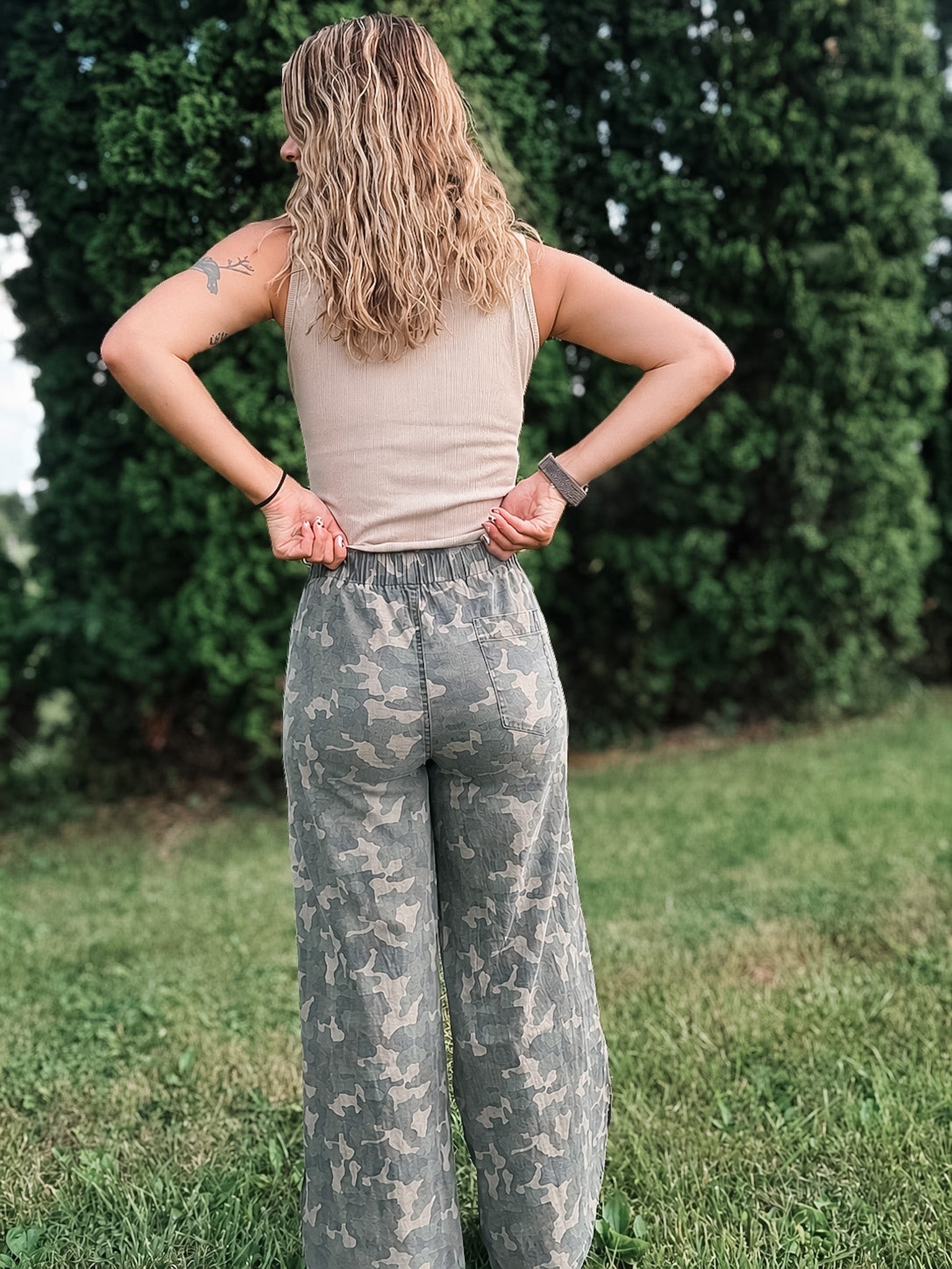 Person wearing camouflage pants and a beige top standing in a grassy area with trees in the background