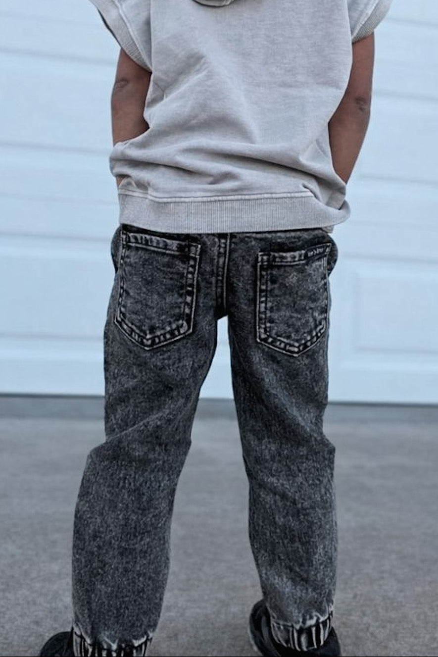 Child wearing a light gray sleeveless hoodie and dark jeans standing in front of a white garage door.