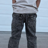 Child wearing a light gray sleeveless hoodie and dark jeans standing in front of a white garage door.