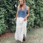 A woman standing outdoors in a grassy area with green hedges, wearing a sleeveless blue denim vest over a white lace midi skirt and black ankle boots.