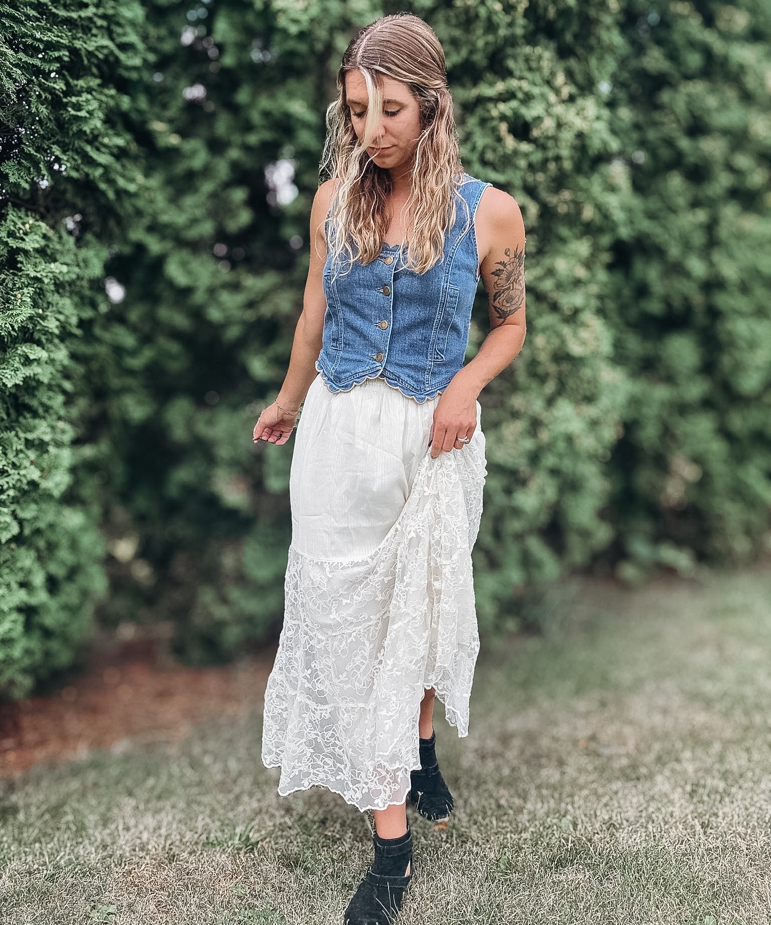 A woman standing outdoors in a grassy area with green hedges, wearing a sleeveless blue denim vest over a white lace midi skirt and black ankle boots.