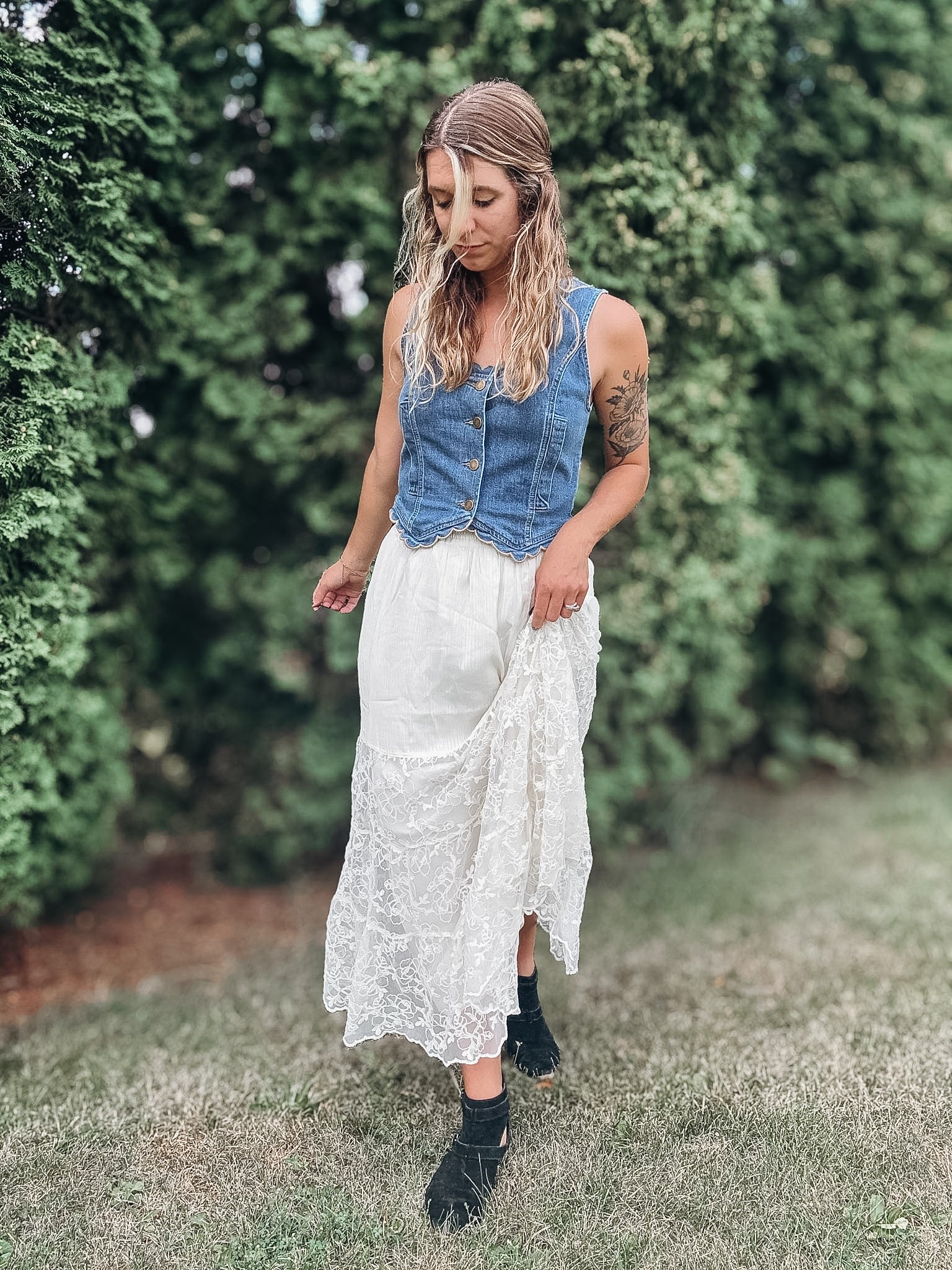 A woman standing outdoors in a grassy area with green hedges, wearing a sleeveless blue denim vest over a white lace midi skirt and black ankle boots.