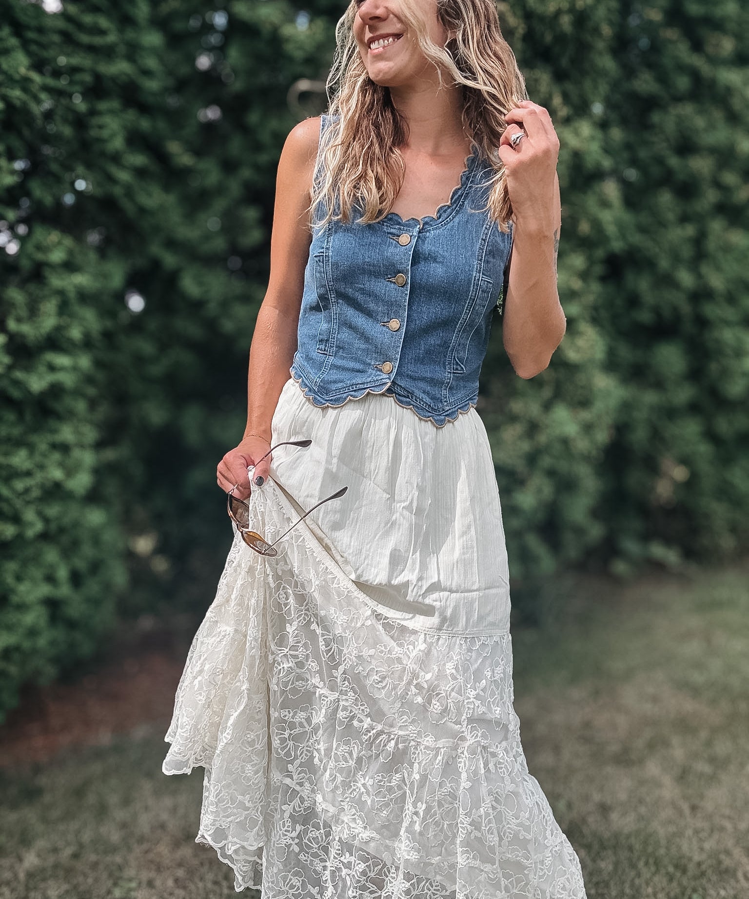 A woman outdoors wearing a sleeveless blue denim vest with buttons over a long white lace maxi skirt, standing in a grassy area with green hedges in the background.
