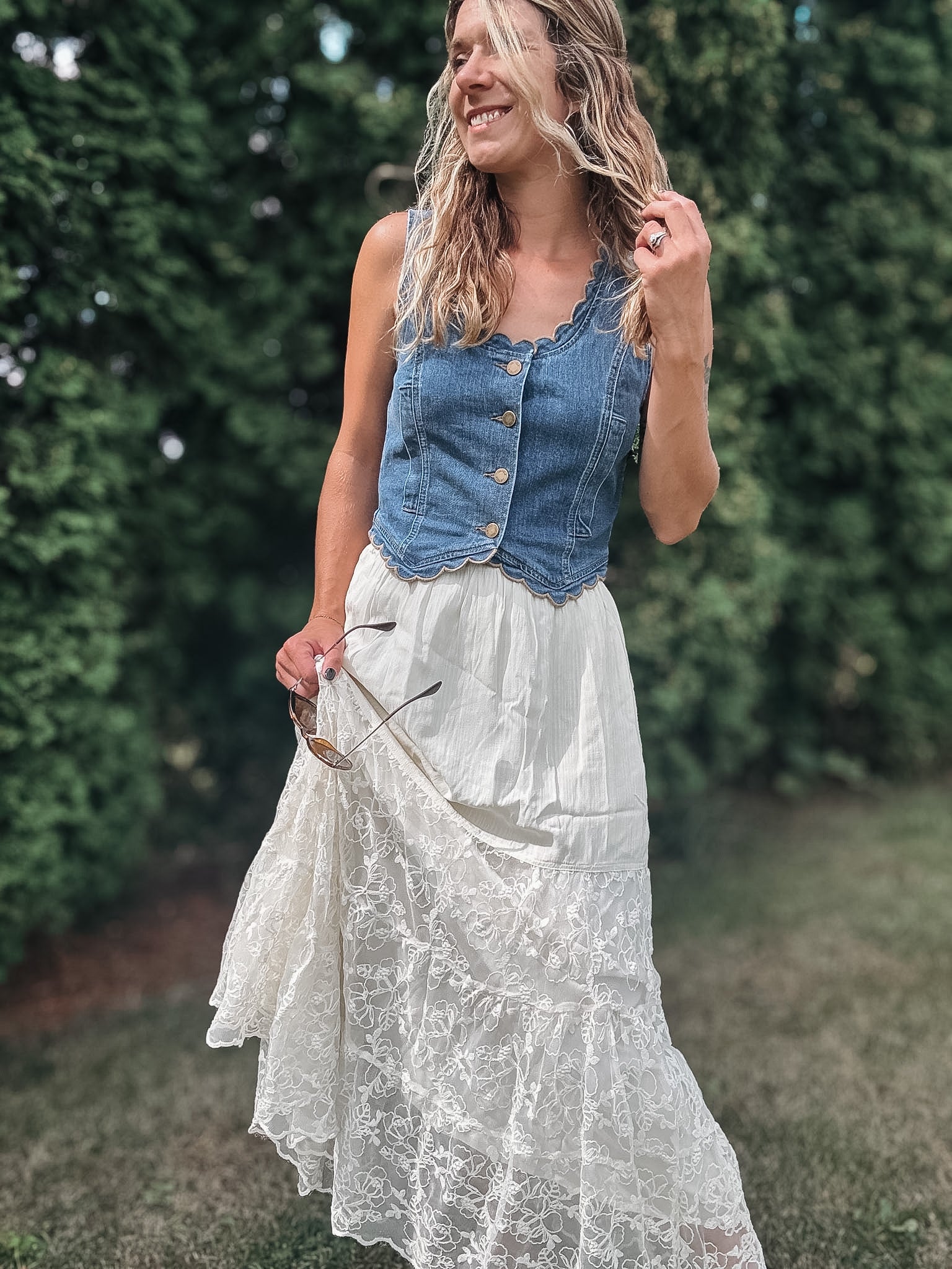 A woman outdoors wearing a sleeveless blue denim vest with buttons over a long white lace maxi skirt, standing in a grassy area with green hedges in the background.