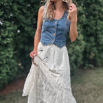 A woman outdoors wearing a sleeveless blue denim vest with buttons over a long white lace maxi skirt, standing in a grassy area with green hedges in the background.
