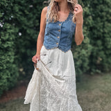 A woman outdoors wearing a sleeveless blue denim vest with buttons over a long white lace maxi skirt, standing in a grassy area with green hedges in the background.