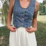 A woman wearing a sleeveless blue denim vest with scalloped edges and front buttons, paired with a white skirt, standing outdoors in a grassy park area.