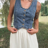 A woman wearing a sleeveless blue denim vest with scalloped edges and front buttons, paired with a white skirt, standing outdoors in a grassy park area.