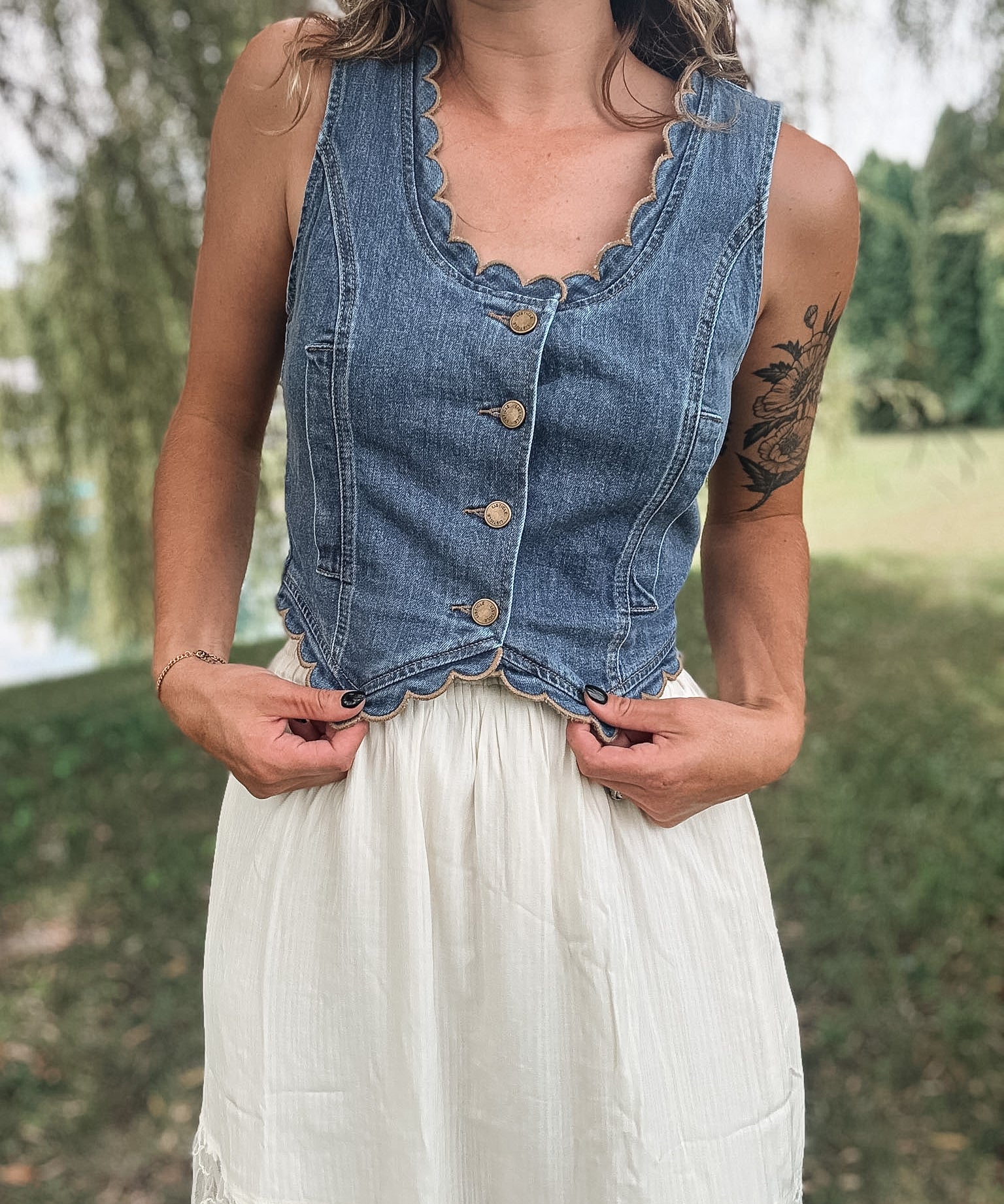 A woman wearing a sleeveless blue denim vest with scalloped edges and front buttons, paired with a white skirt, standing outdoors in a grassy park area.