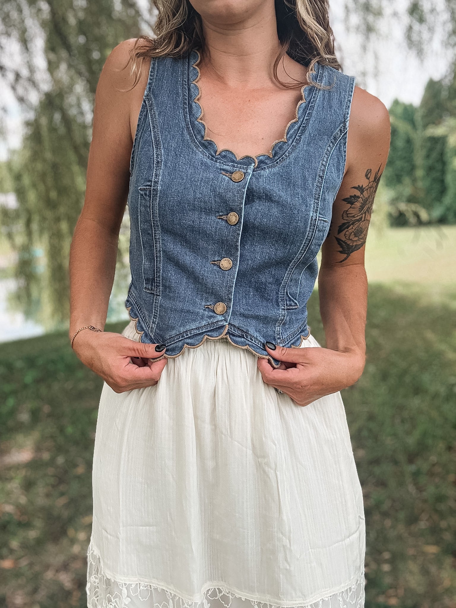 A woman wearing a sleeveless blue denim vest with scalloped edges and front buttons, paired with a white skirt, standing outdoors in a grassy park area.