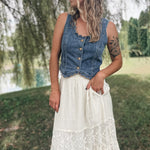A woman standing outdoors in a grassy park area wearing a sleeveless blue denim vest with buttons and a long white lace maxi skirt.