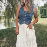 A woman standing outdoors in a grassy park area wearing a sleeveless blue denim vest with buttons and a long white lace maxi skirt.