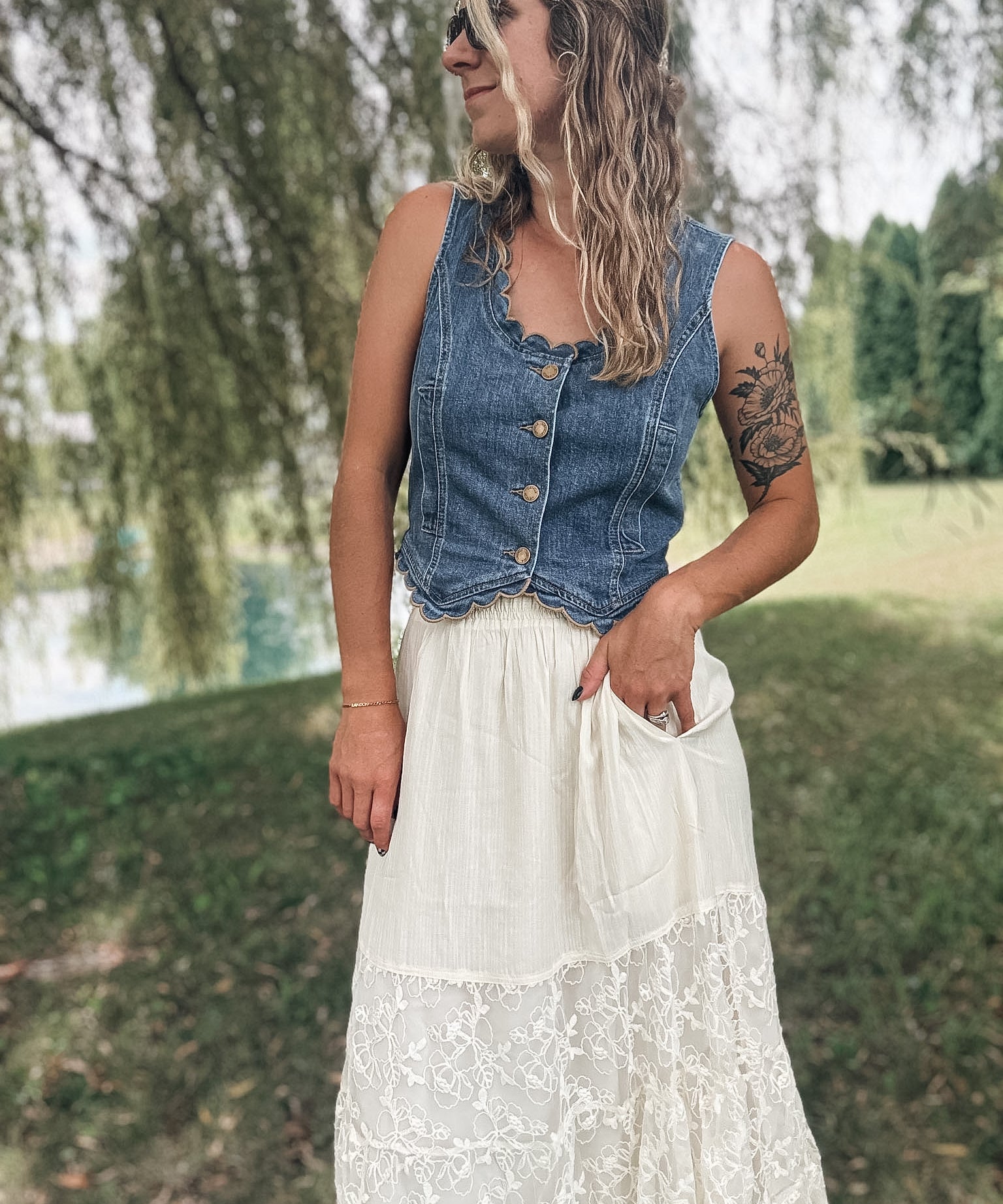 A woman standing outdoors in a grassy park area wearing a sleeveless blue denim vest with buttons and a long white lace maxi skirt.