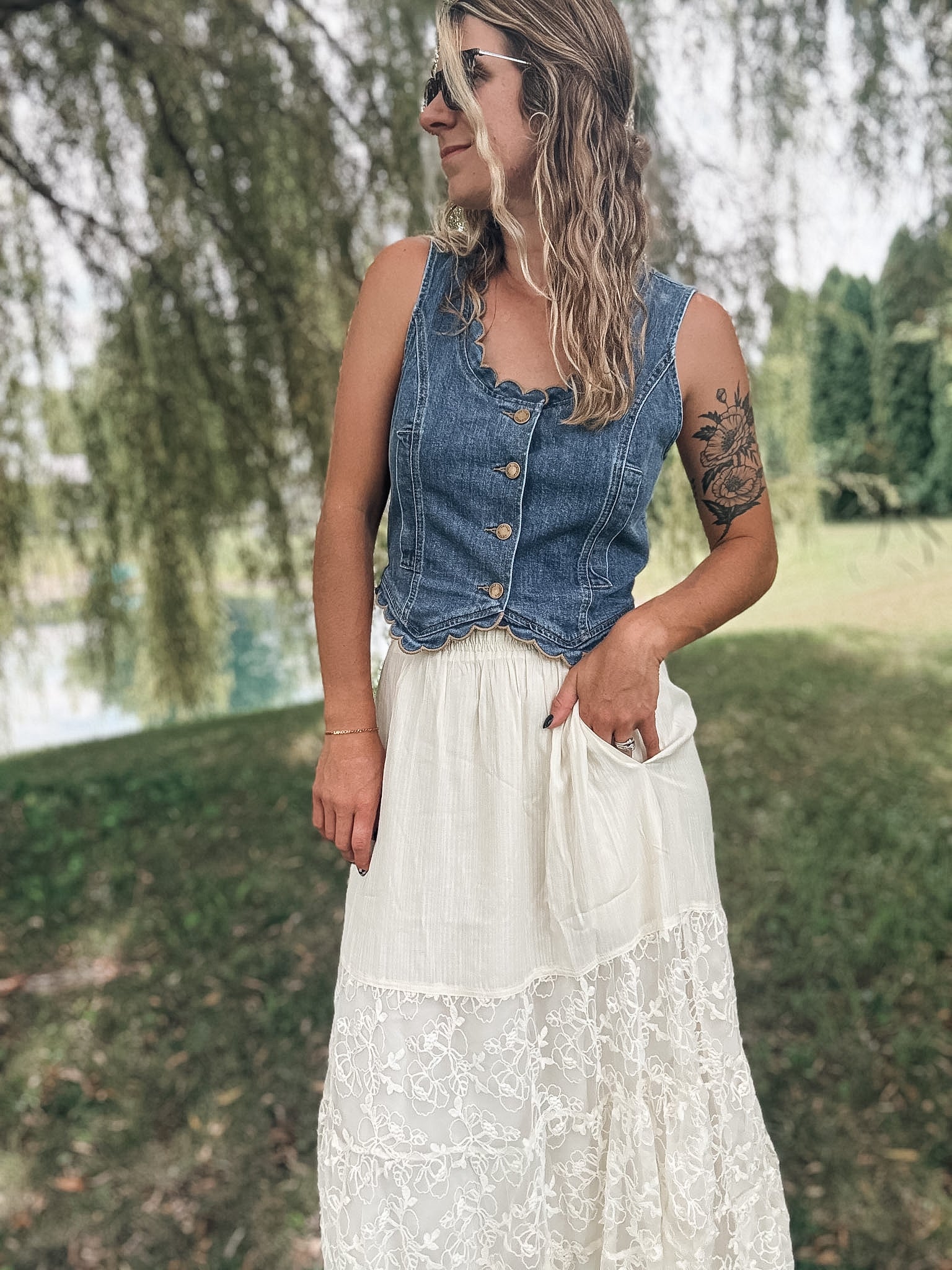 A woman standing outdoors in a grassy park area wearing a sleeveless blue denim vest with buttons and a long white lace maxi skirt.