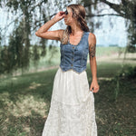A woman standing outdoors in a grassy field under trees, wearing a sleeveless denim vest over a white lace maxi skirt.