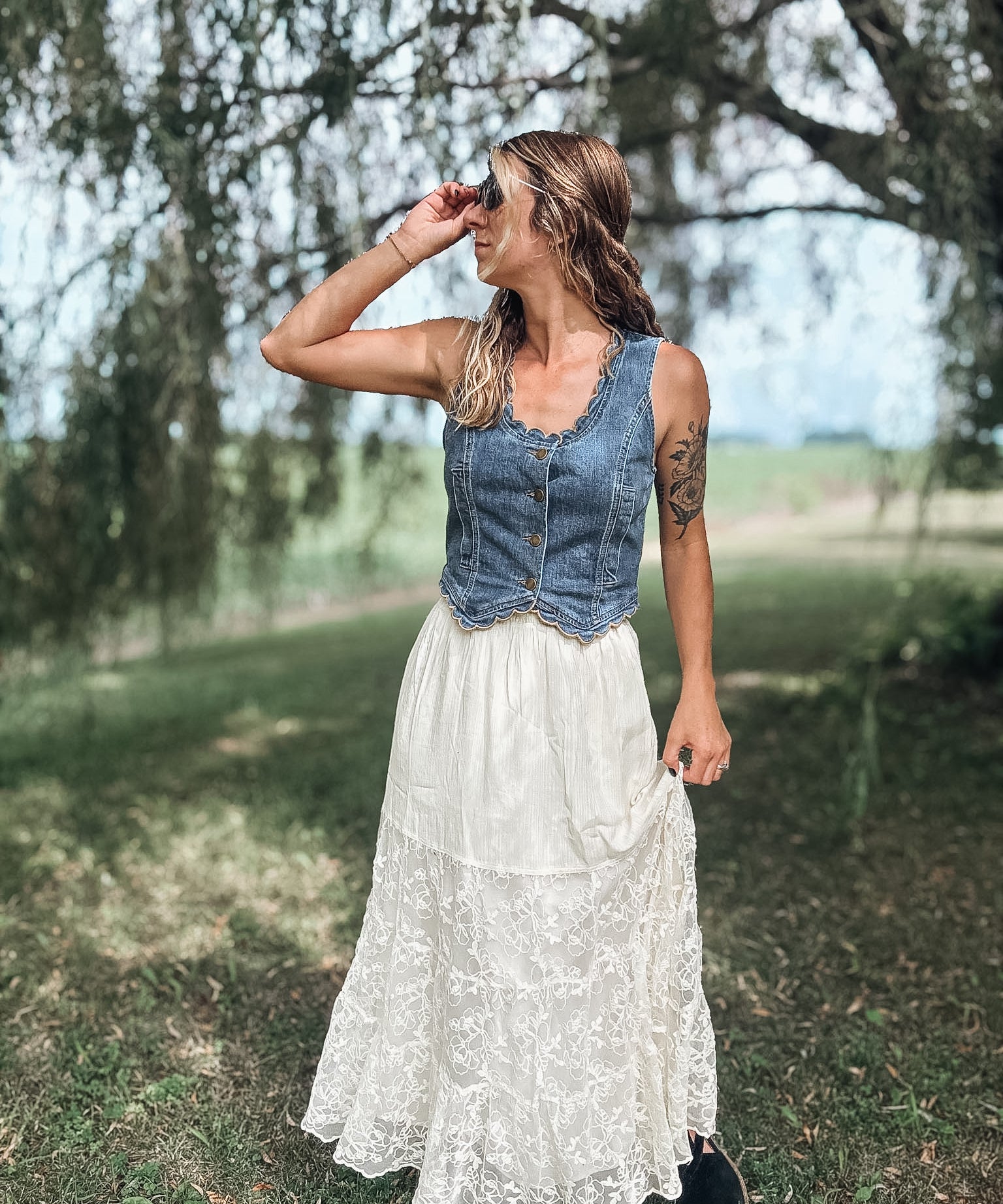 A woman standing outdoors in a grassy field under trees, wearing a sleeveless denim vest over a white lace maxi skirt.