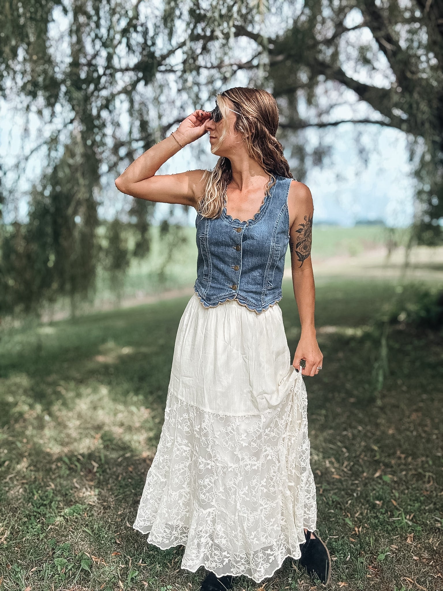 A woman standing outdoors in a grassy field under trees, wearing a sleeveless denim vest over a white lace maxi skirt.